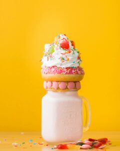 Vibrant milkshake adorned with a donut, whipped cream, and candy against a yellow background.