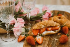 Close-up of delicious croissants with strawberries and cream, accompanied by pink flowers and wine glasses.