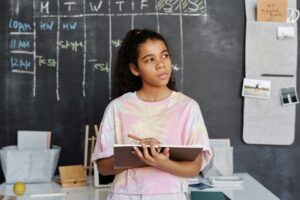 A teenager in a pink shirt ponders while holding a notebook in a classroom setting.