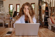 A thoughtful woman working remotely on a laptop in a cozy indoor setting.